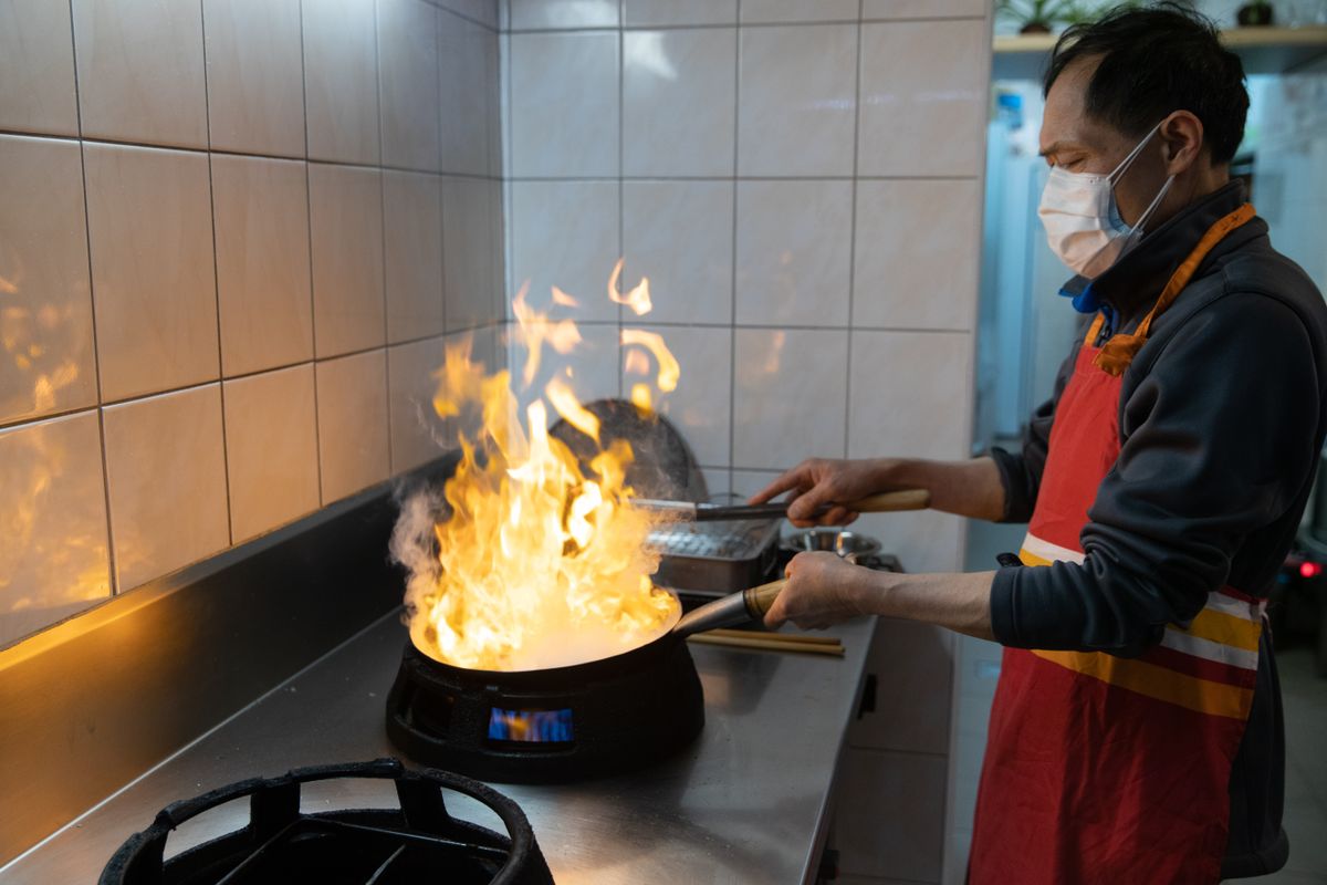 An Asian chef wearing a face mask cooks a flaming dish in a commercial kitchen.