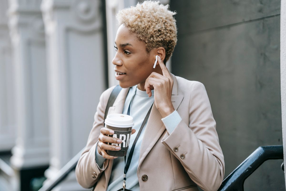 Cheerful young African American female student in classy jacket listening to music via wireless earbuds and drinking coffee near railing