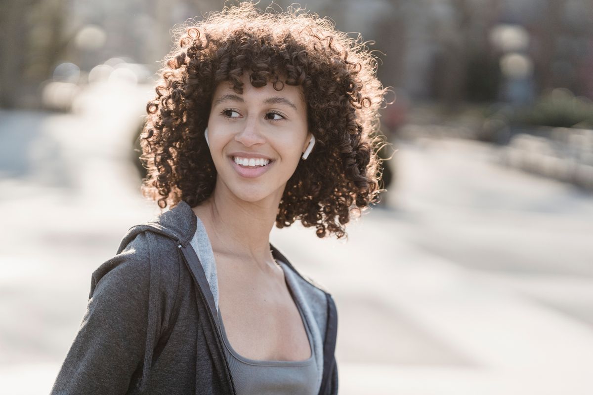 Cheerful young ethnic female athlete with curly hair listening to music from earbuds while looking away in town in back lit