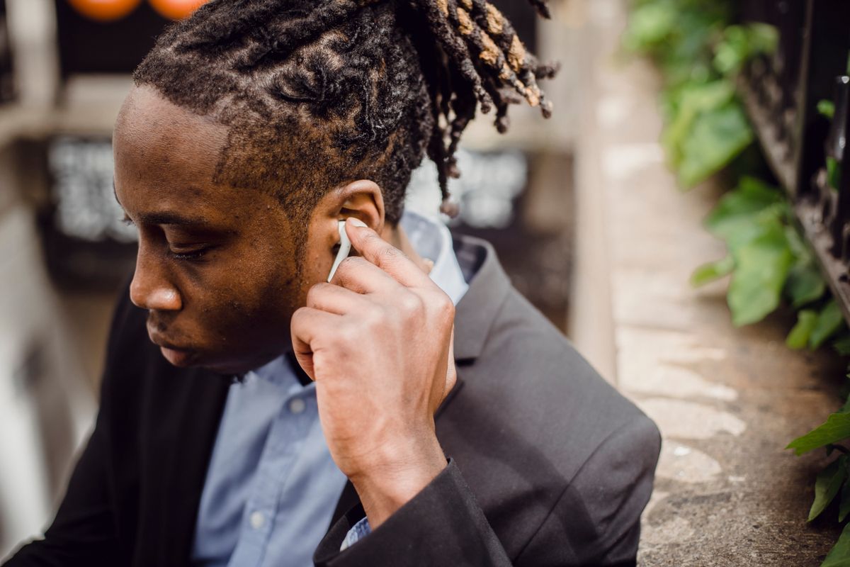 High angle of crop young African American man in smart suit with dreadlocks in ponytail listening to music through wireless earphones while standing on street on sunny day