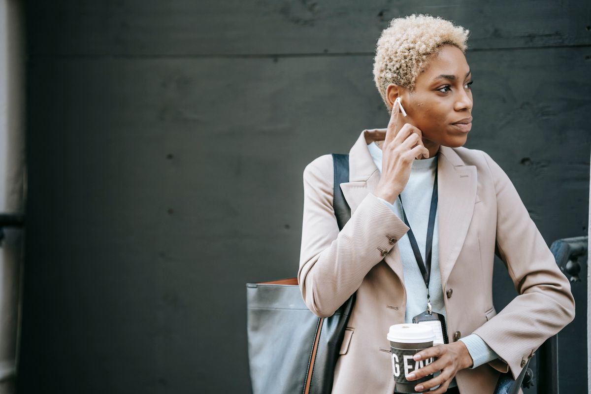 Young pondering black female manager with beverage to go and wireless earbud listening to music while looking away on street