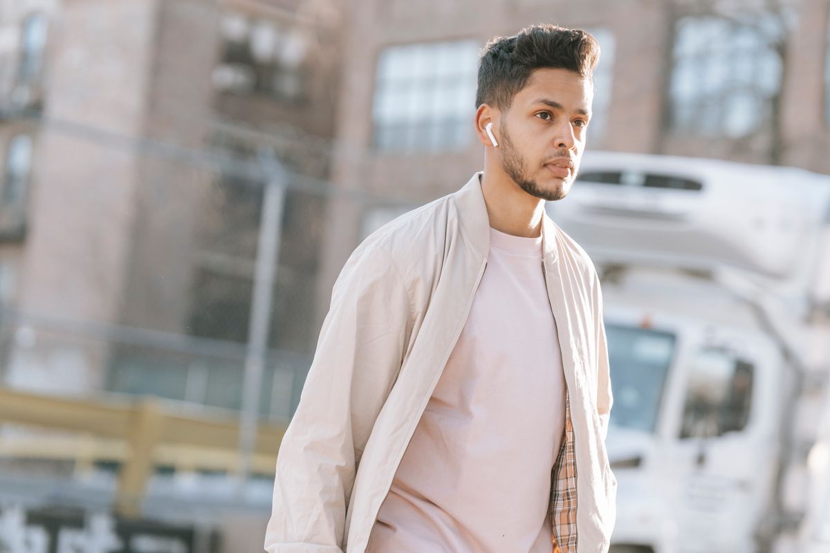 Masculine ethnic male with modern haircut listening to music from wireless earbud while looking away in sunny city