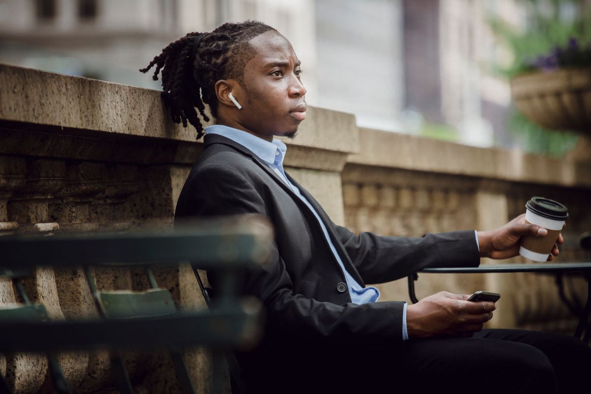 Side view of concentrated young African American man with dreadlocks in elegant suit listening to music in true wireless earphones while drinking takeaway coffee on terrace of modern building