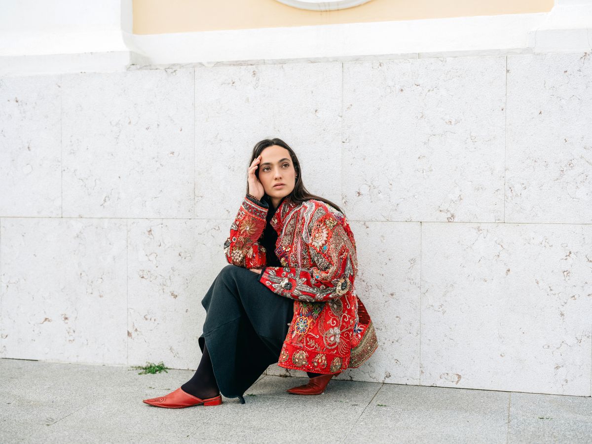 A stylish woman crouches against a marble wall in Lisbon, showcasing modern fashion with a vibrant red jacket.
