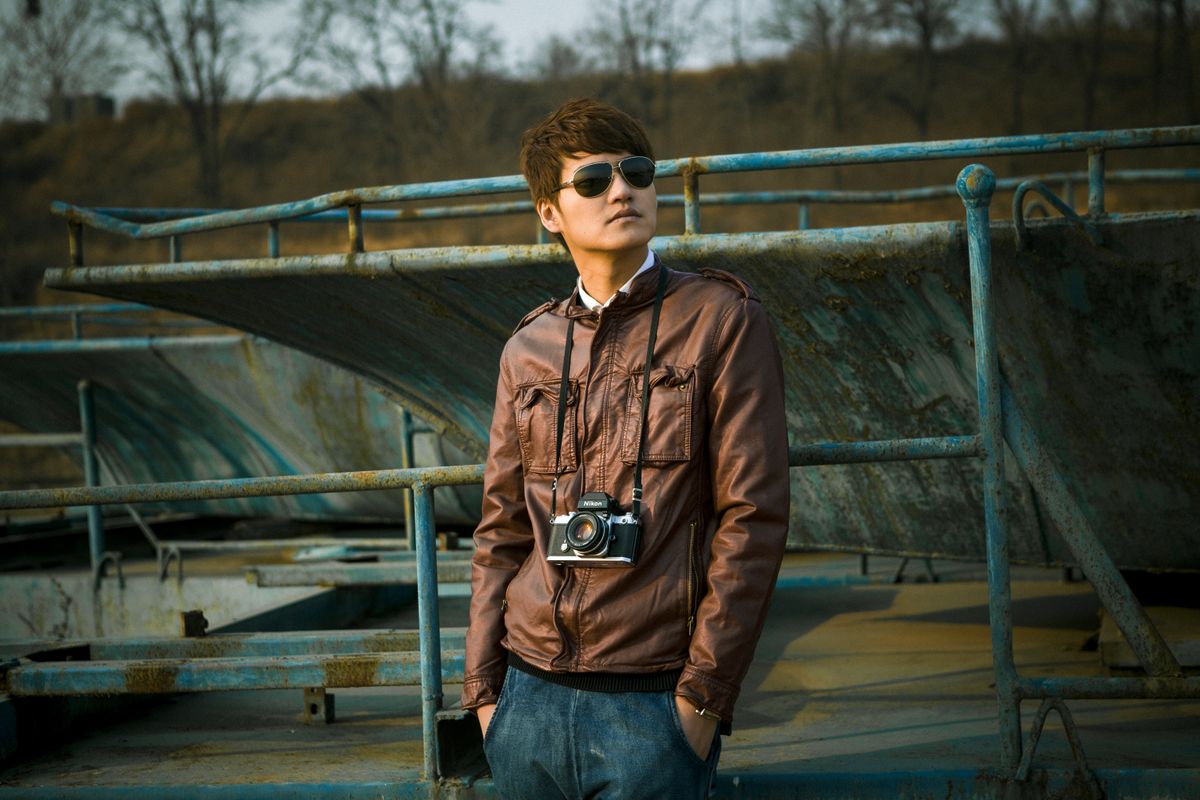Stylish man wearing sunglasses and leather jacket holding a vintage camera in an outdoor setting.