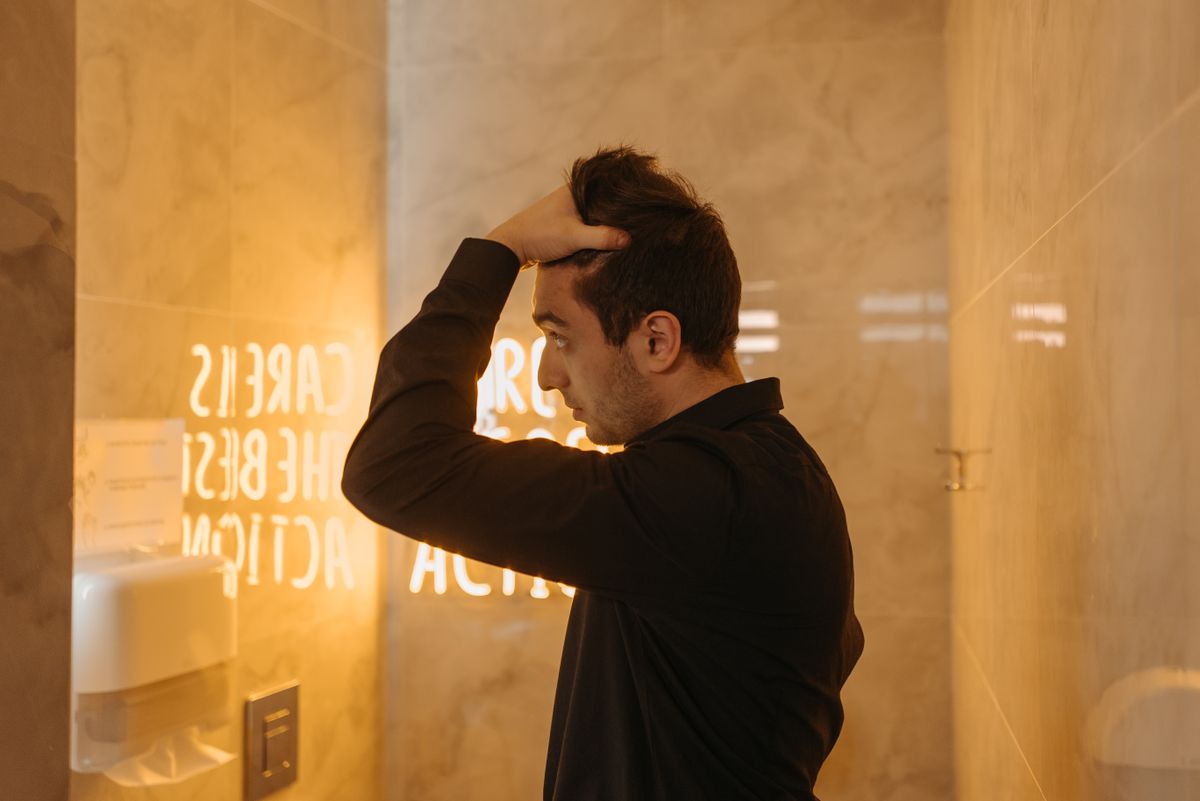 Man in black sleeves standing in a bathroom, looking thoughtful with hand on forehead.