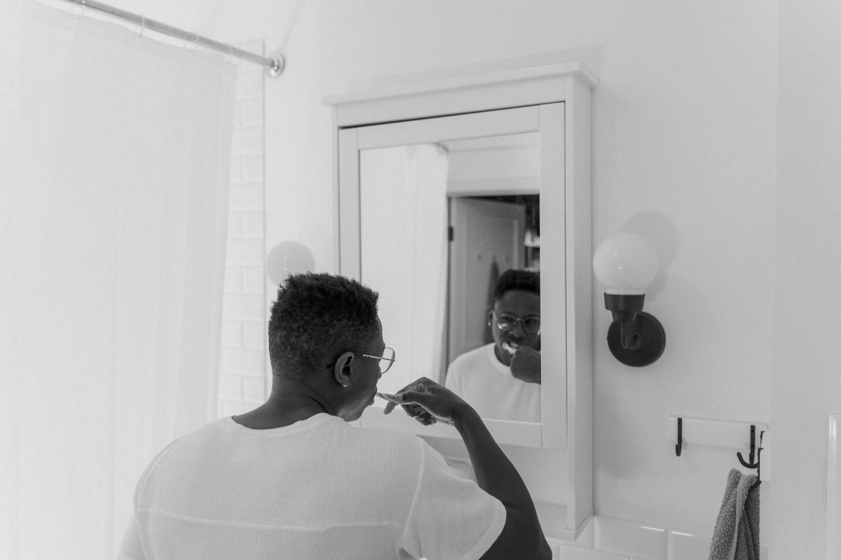 A black and white photo of a man brushing his teeth in front of a bathroom mirror.
