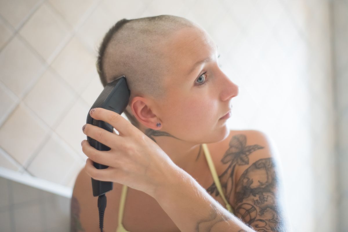 A woman with tattoos shaving her head with an electric razor in a well-lit indoor setting.