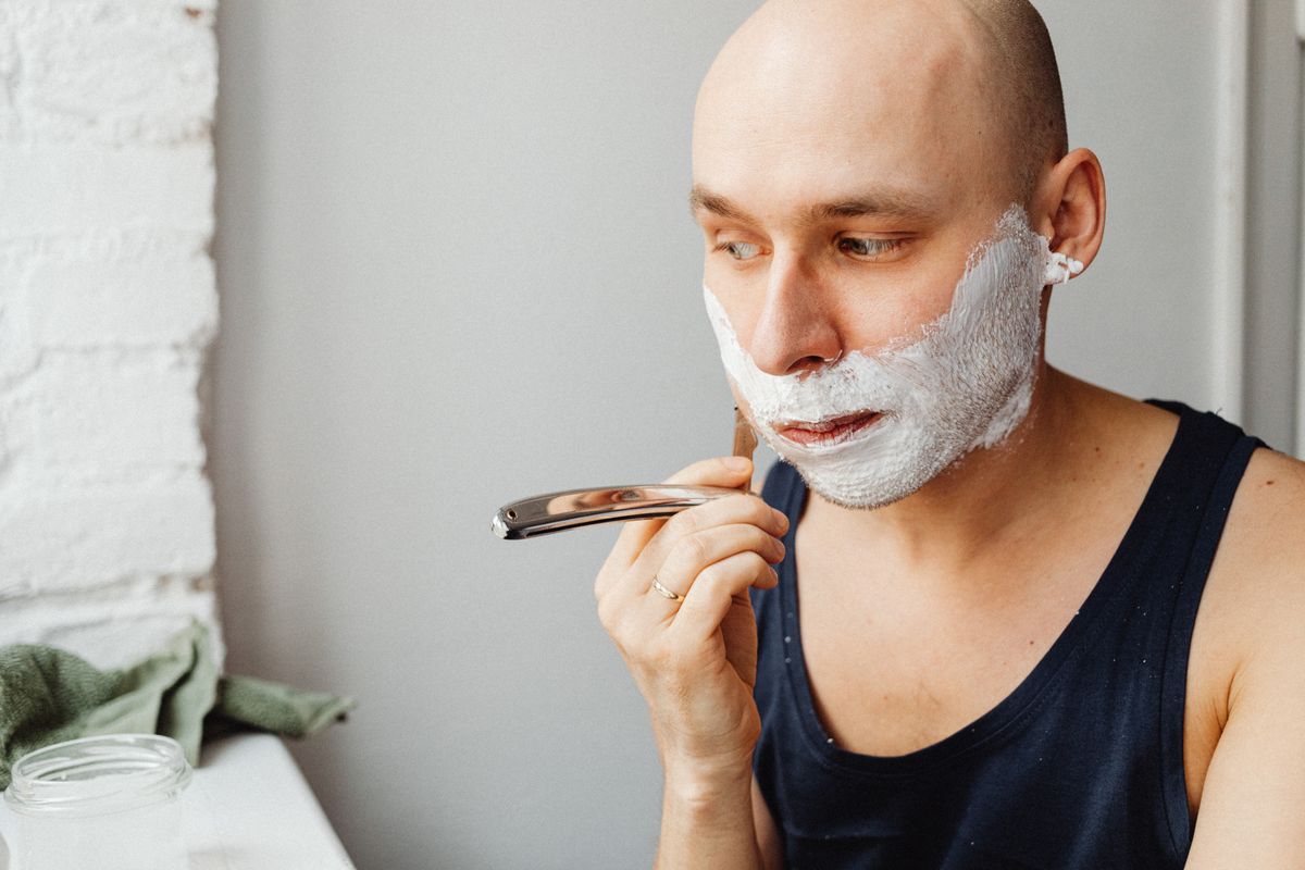 Adult bald man applying shaving cream and using a straight razor indoors.