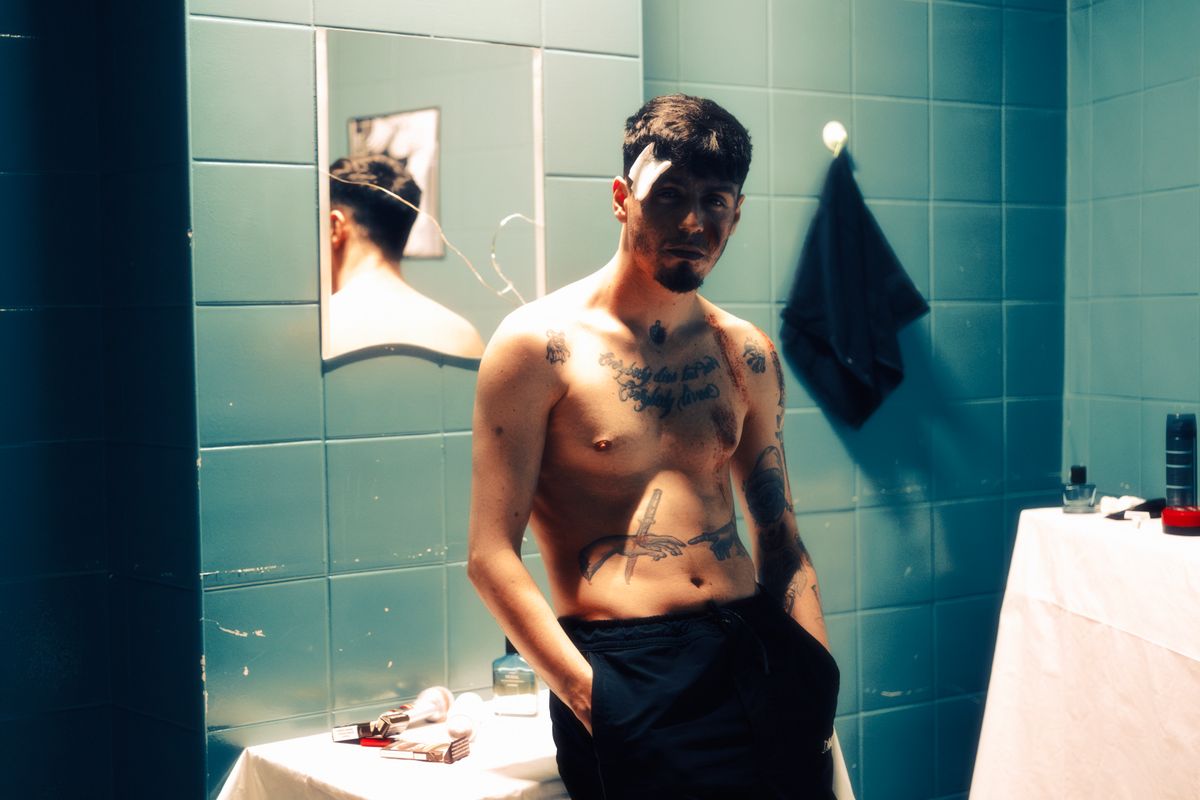 A shirtless, tattooed man stands in a dimly lit bathroom, radiating mystery.