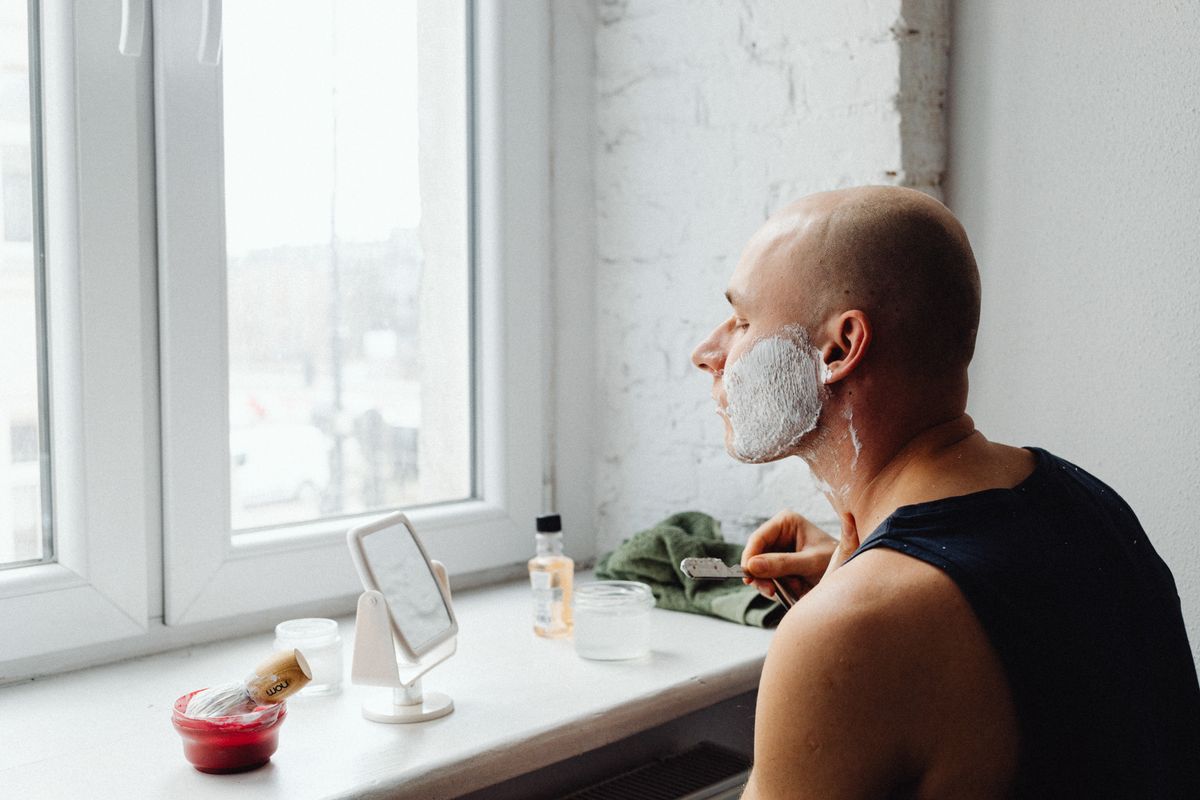 Bald man applying shaving foam with brush indoors by window, morning routine.