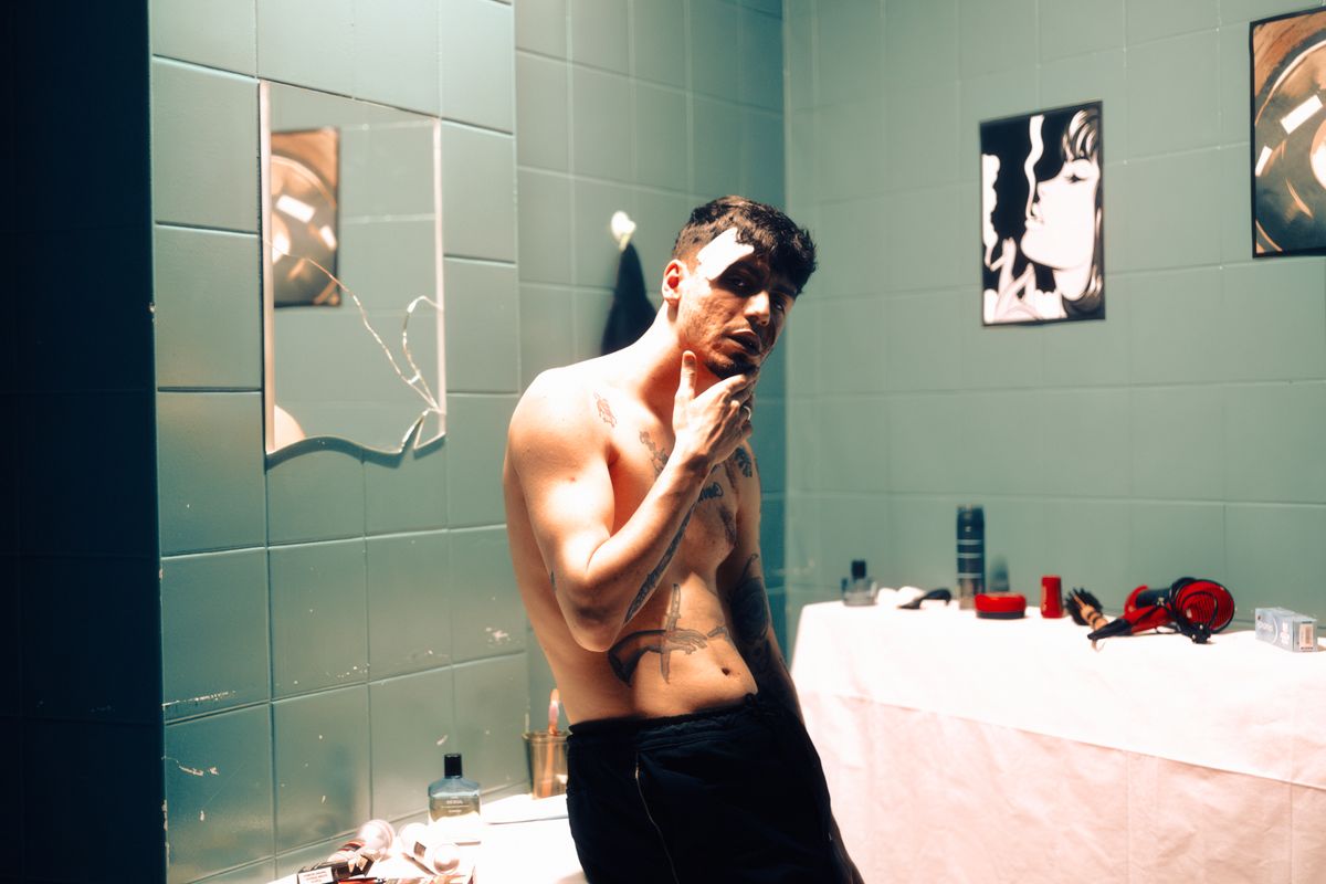 A tattooed man poses thoughtfully in a dimly lit bathroom with art on the walls.