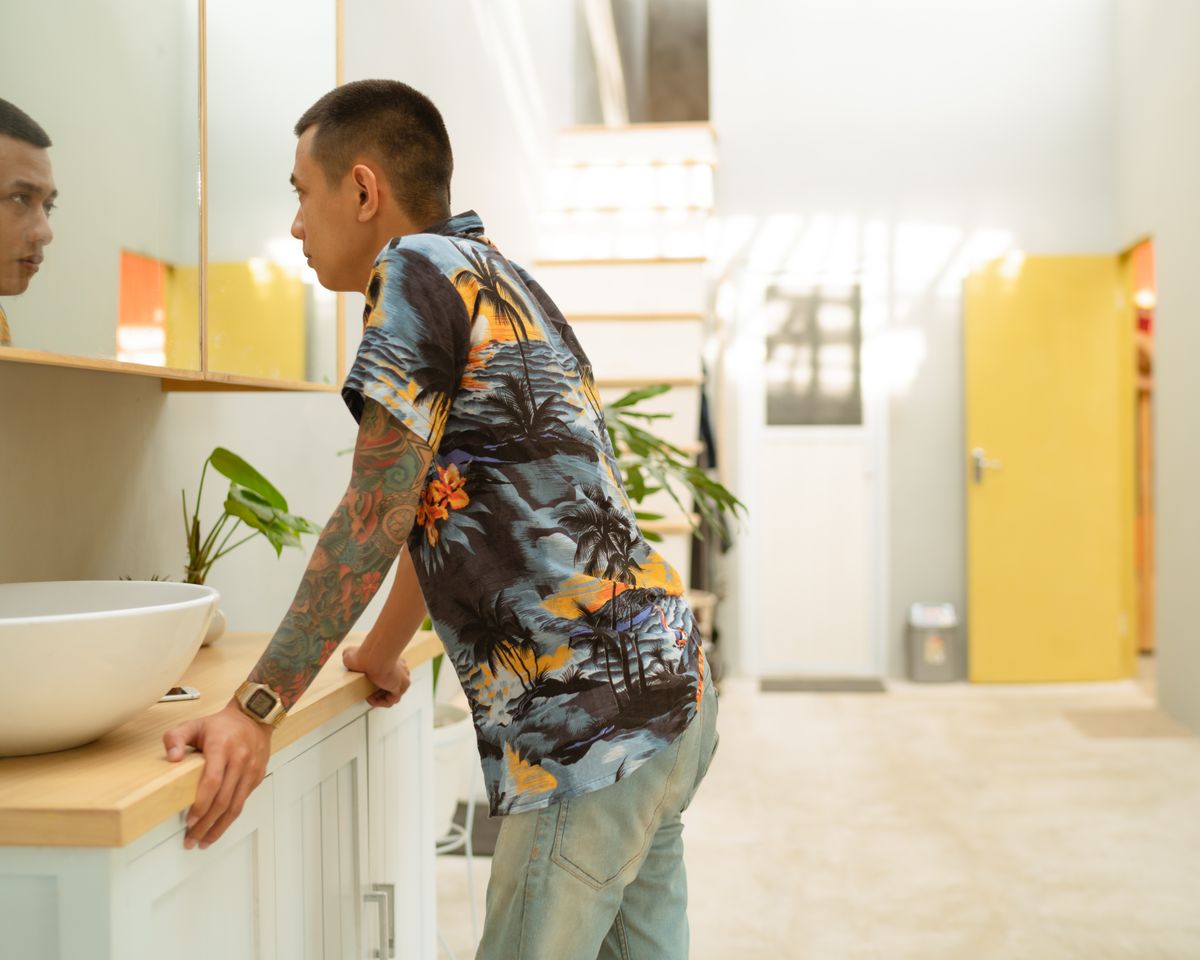 Man in a colorful Hawaiian shirt with tattoo, looking into a mirror in a bright, modern bathroom.