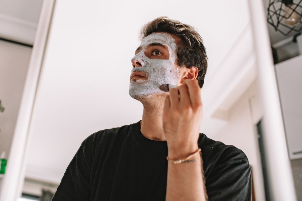 Adult man applying a facial mask while looking in the mirror at home, promoting skincare routine.