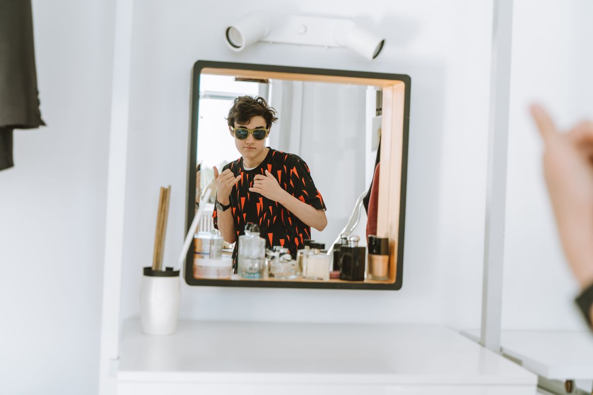 A stylish young man wearing sunglasses poses confidently in front of a mirror indoors.