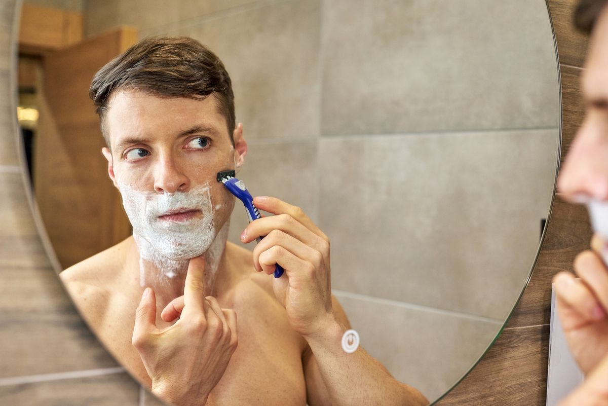 A man uses a razor for shaving in front of a bathroom mirror, focusing carefully.