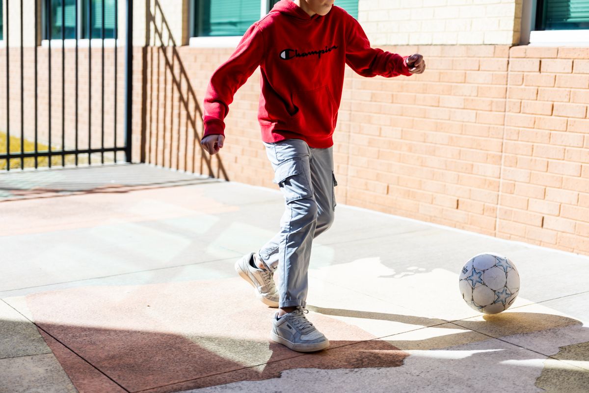 A teenager in a red hoodie playing soccer on a sunny day in a courtyard.