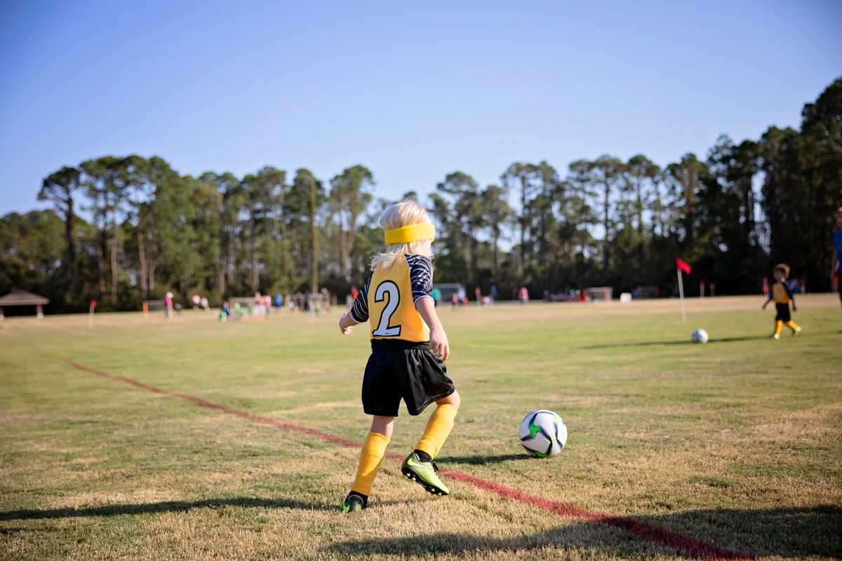 A young soccer player dribbles the ball on a sunny field surrounded by trees.