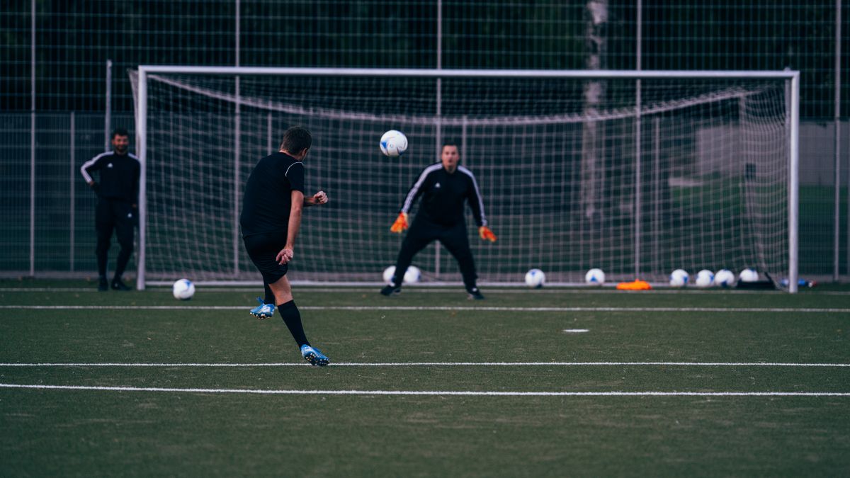Male football player kicking ball towards goalkeeper standing against goal during training in field