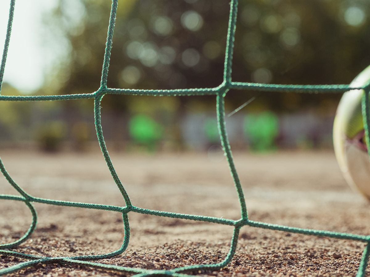 Ground level of green soccer net on rough terrain near ball on blurred background