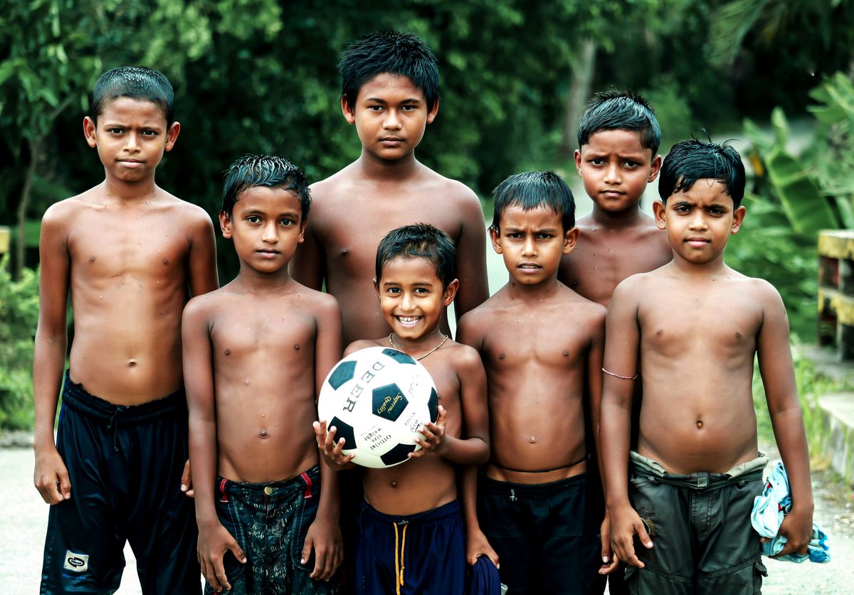 A group of six South Asian boys, standing outdoors, with one holding a soccer ball.
