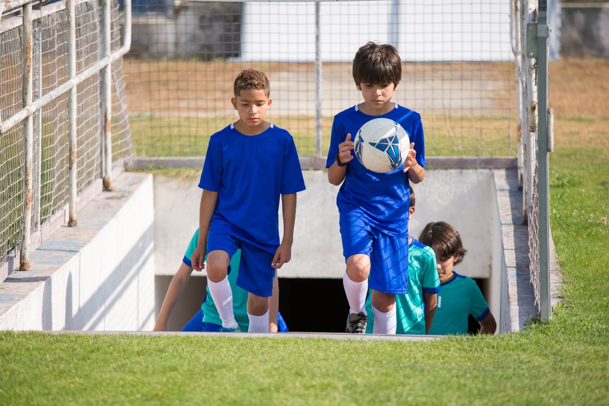 Children walking onto a soccer field during a sunny day, holding a soccer ball.