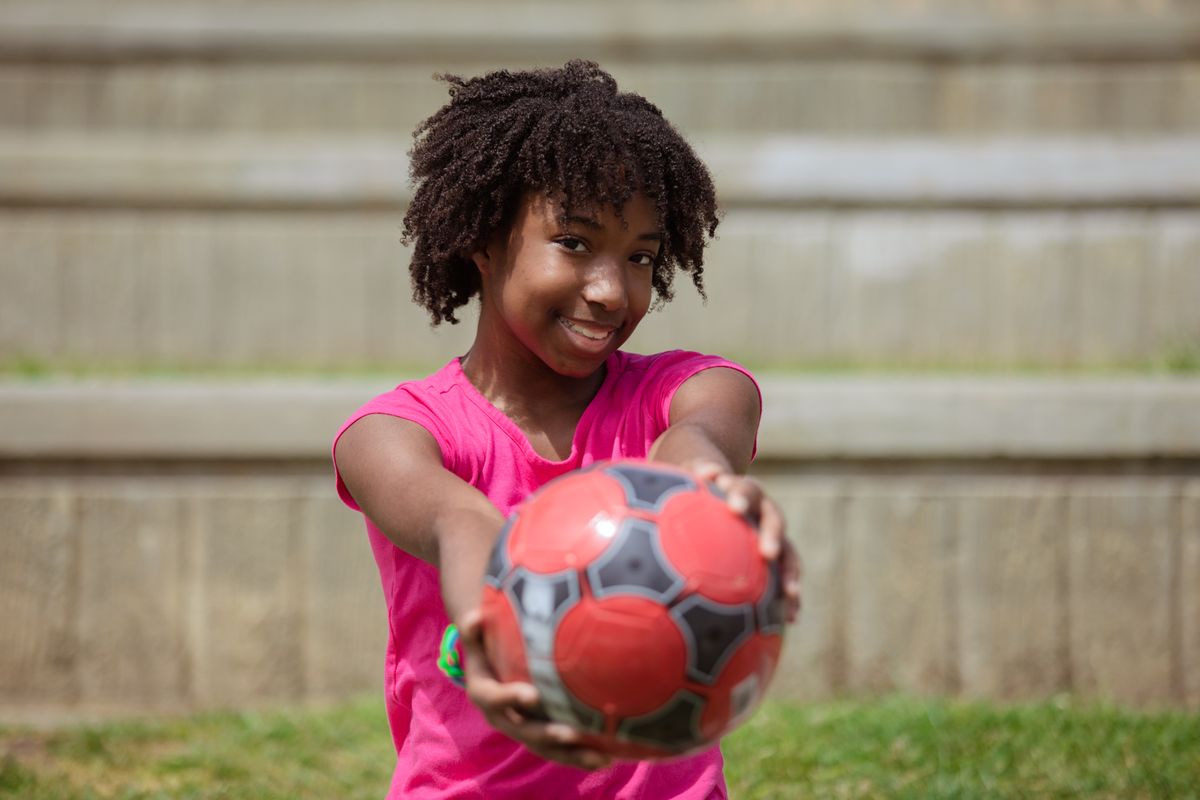 A joyful young girl in a pink shirt holding a soccer ball with a vibrant smile.