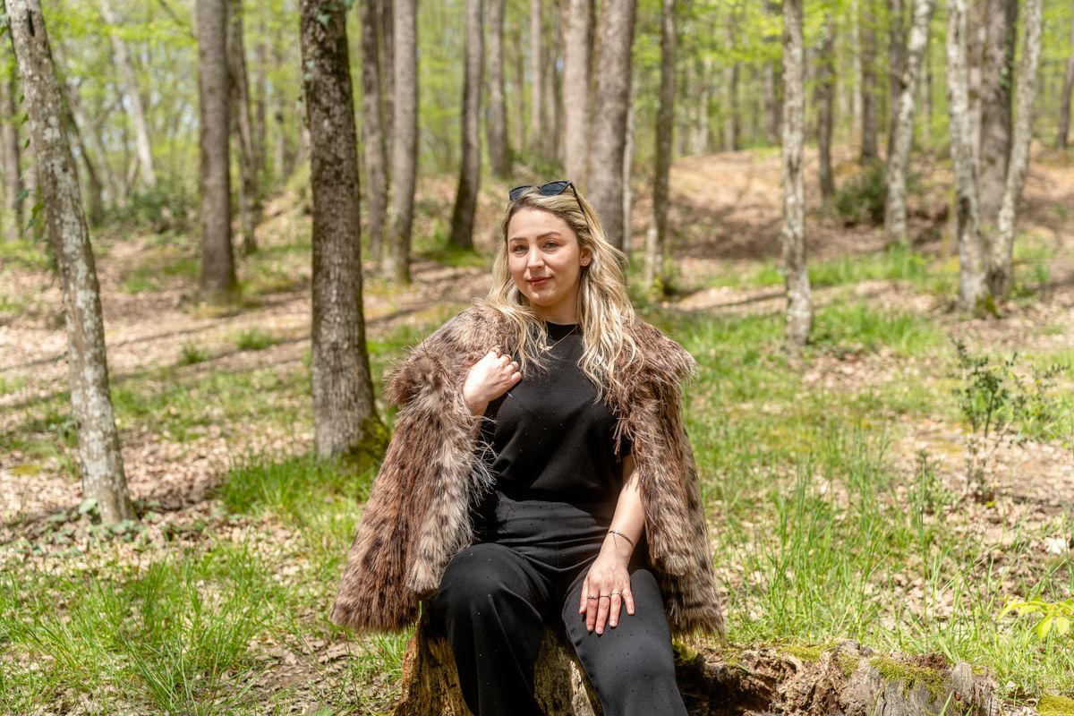 Portrait of a woman sitting on a tree stump in a sunlit forest, wearing a faux fur coat.