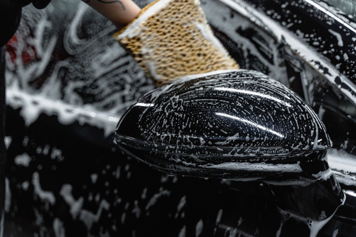 Hand scrubbing a car mirror with a sponge covered in soap at a car wash.
