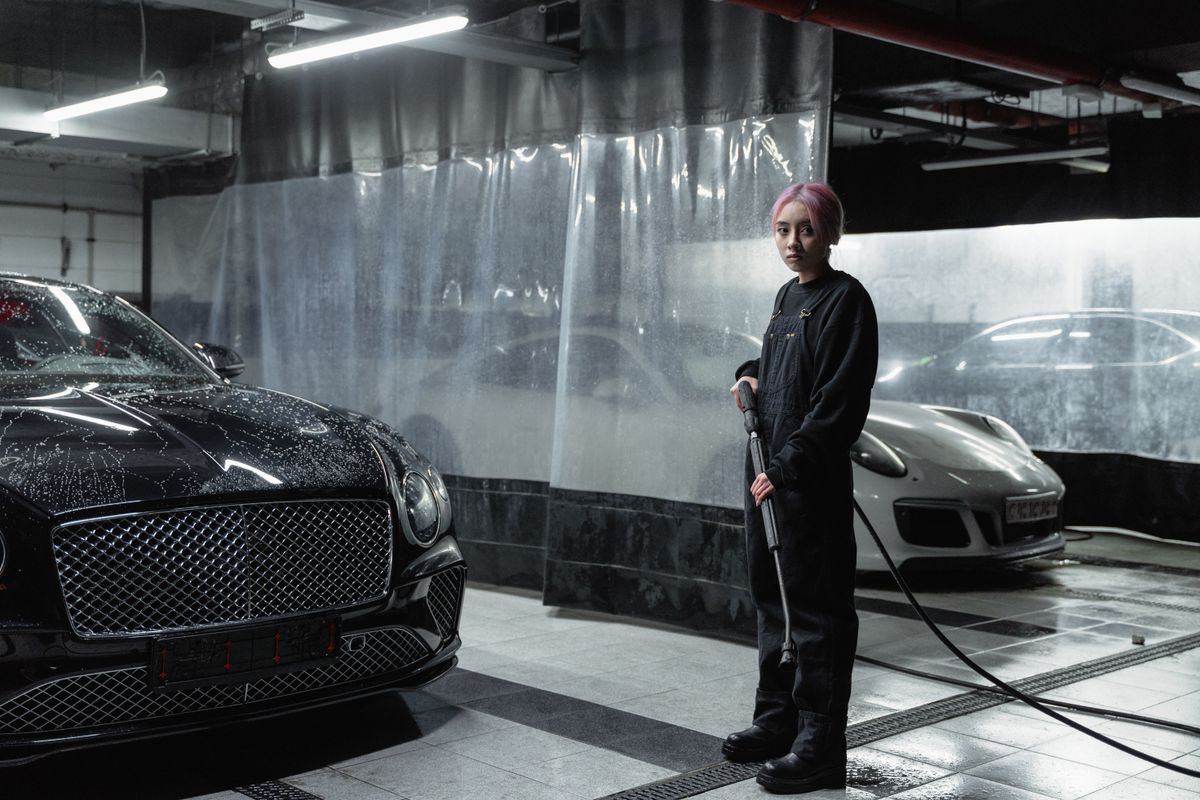 A woman with pink hair operates a power spray in a carwash garage with luxury cars.