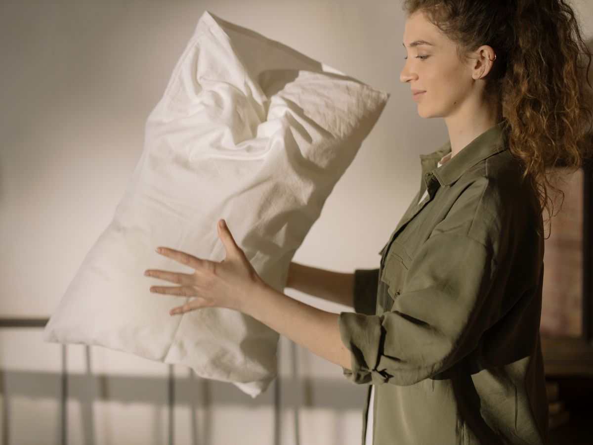 A woman in a casual shirt fluffing a pillow in a cozy, well-lit bedroom.