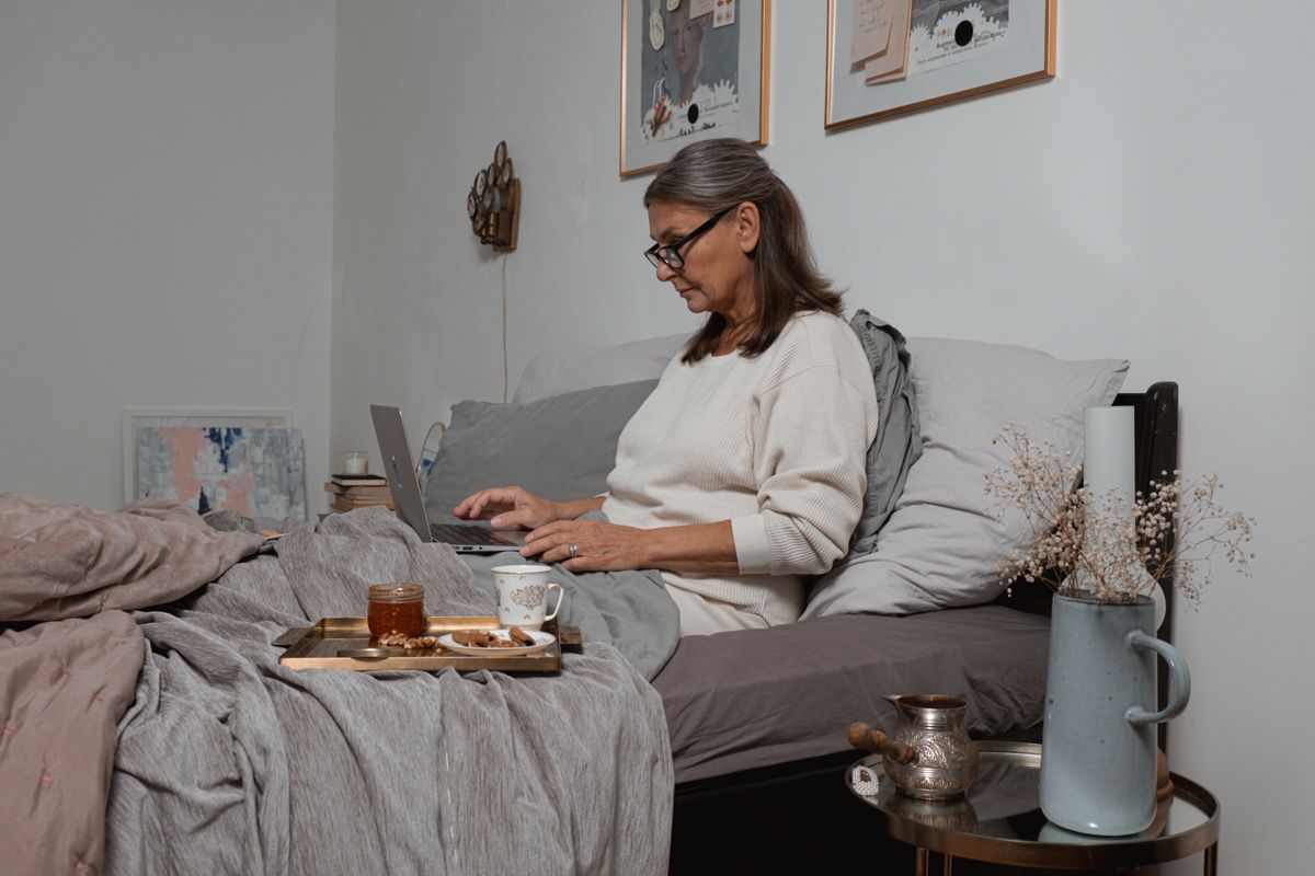 Elderly woman in cozy setting using a laptop in bed with coffee and snacks.