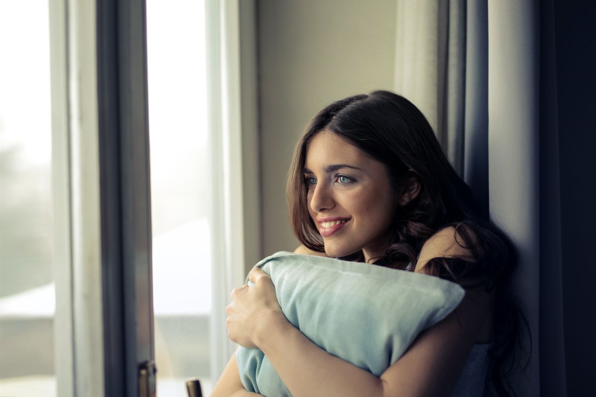 A woman sits comfortably by a window, holding a pillow and smiling happily indoors.