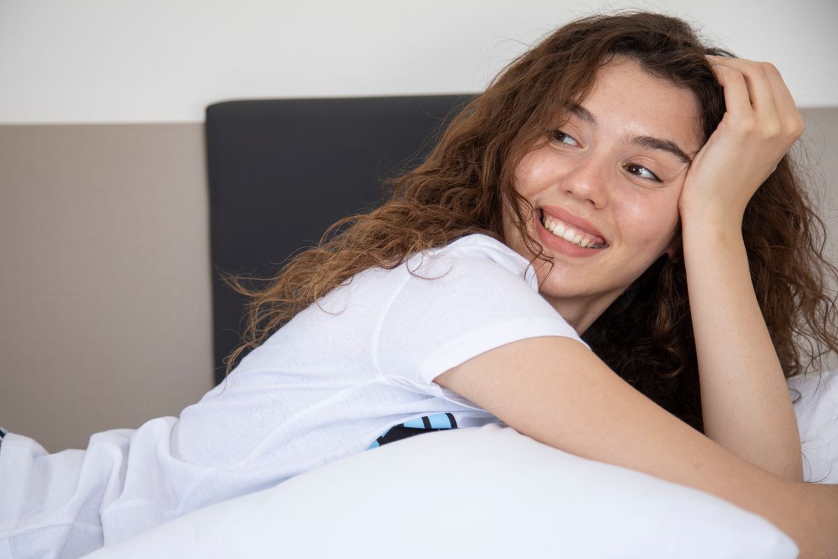 A young woman with curly hair smiles while lying on her bed, creating a cozy and relaxed vibe.