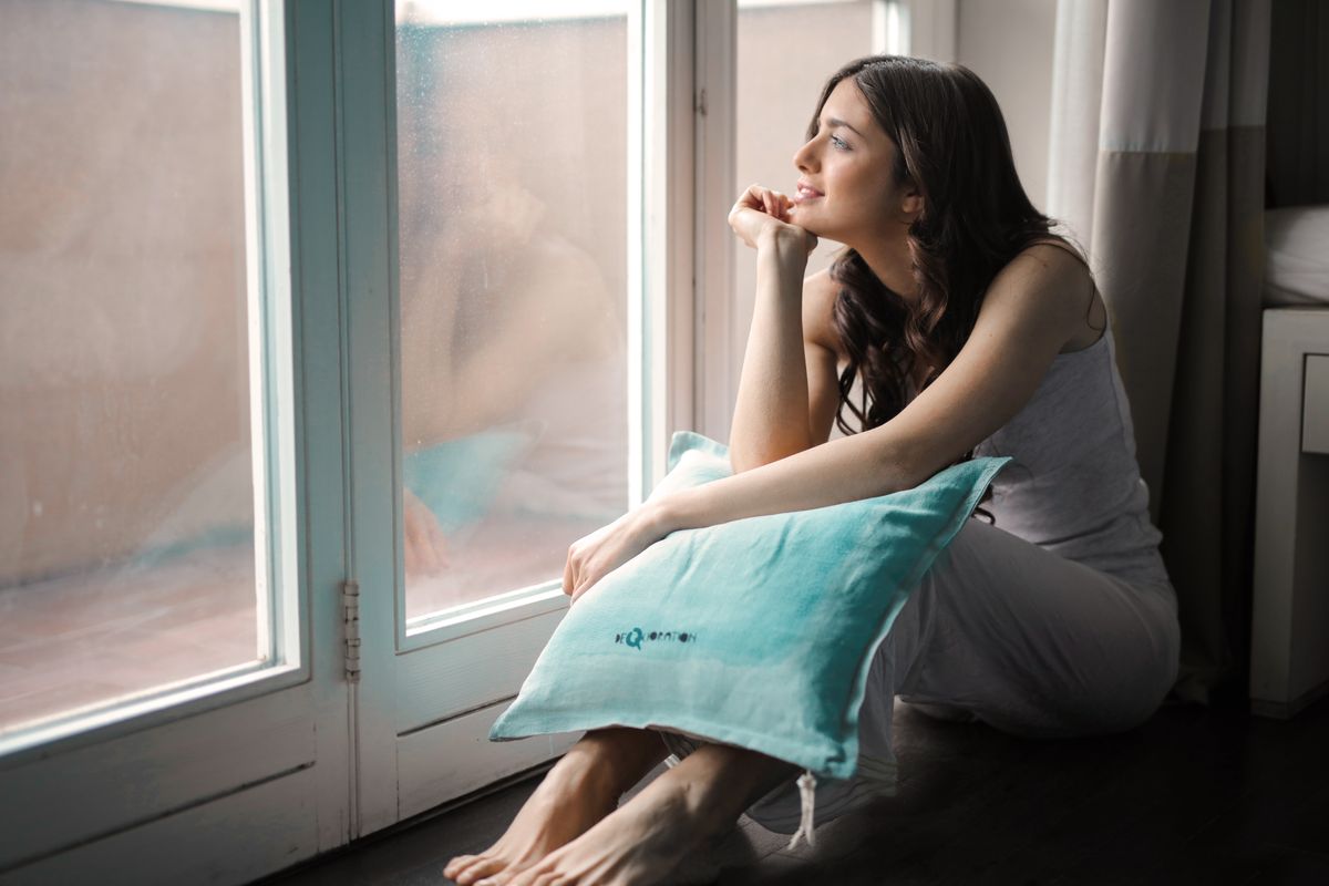 A thoughtful woman gazing outside while sitting by a window with a pillow.