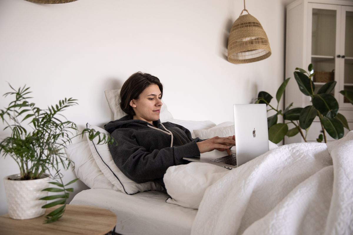 Woman lying comfortably in bed with a laptop, surrounded by cozy interior and plants.