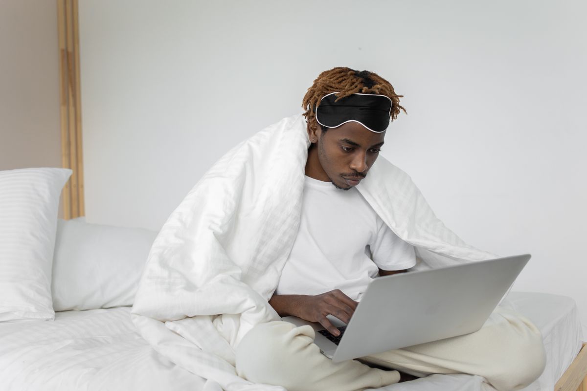 A young man with dreadlocks sits on a bed working on his laptop in a minimalist bedroom.