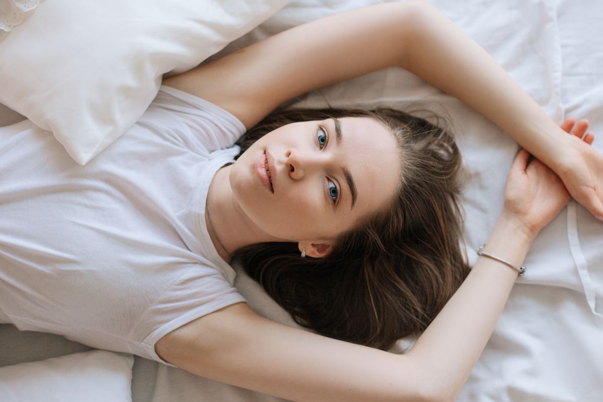 A woman lying down on her bed with white linens, in a relaxed pose wearing a white shirt.