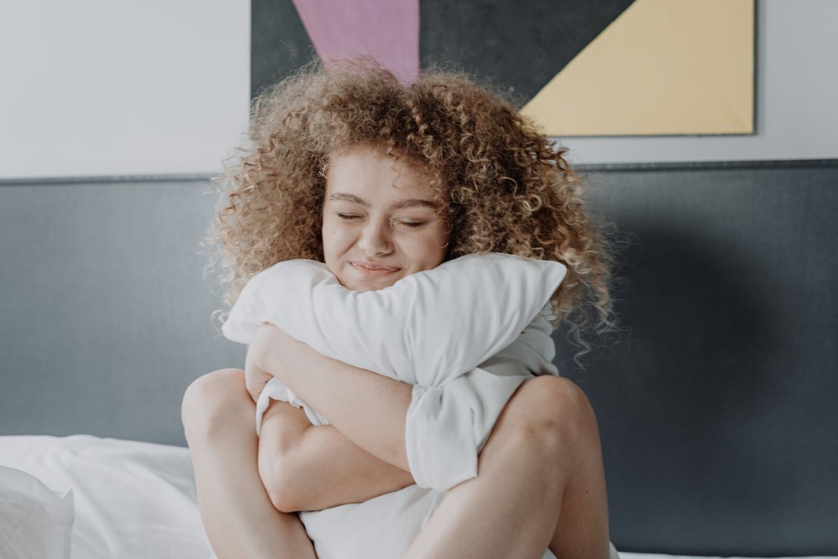 A woman with curly hair enjoys a cozy morning, embracing a pillow in white bed linen.