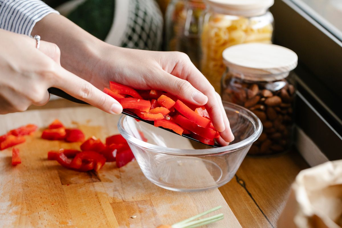 Unrecognizable female cook with knife putting cut red bell pepper from cutting board into glass bowl while cooking in kitchen