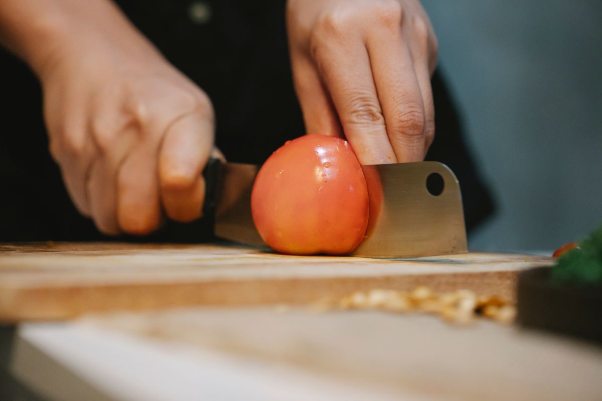 Crop unrecognizable chef slicing ripe wet red tomato with knife on wooden cutting board for cooking healthy dish