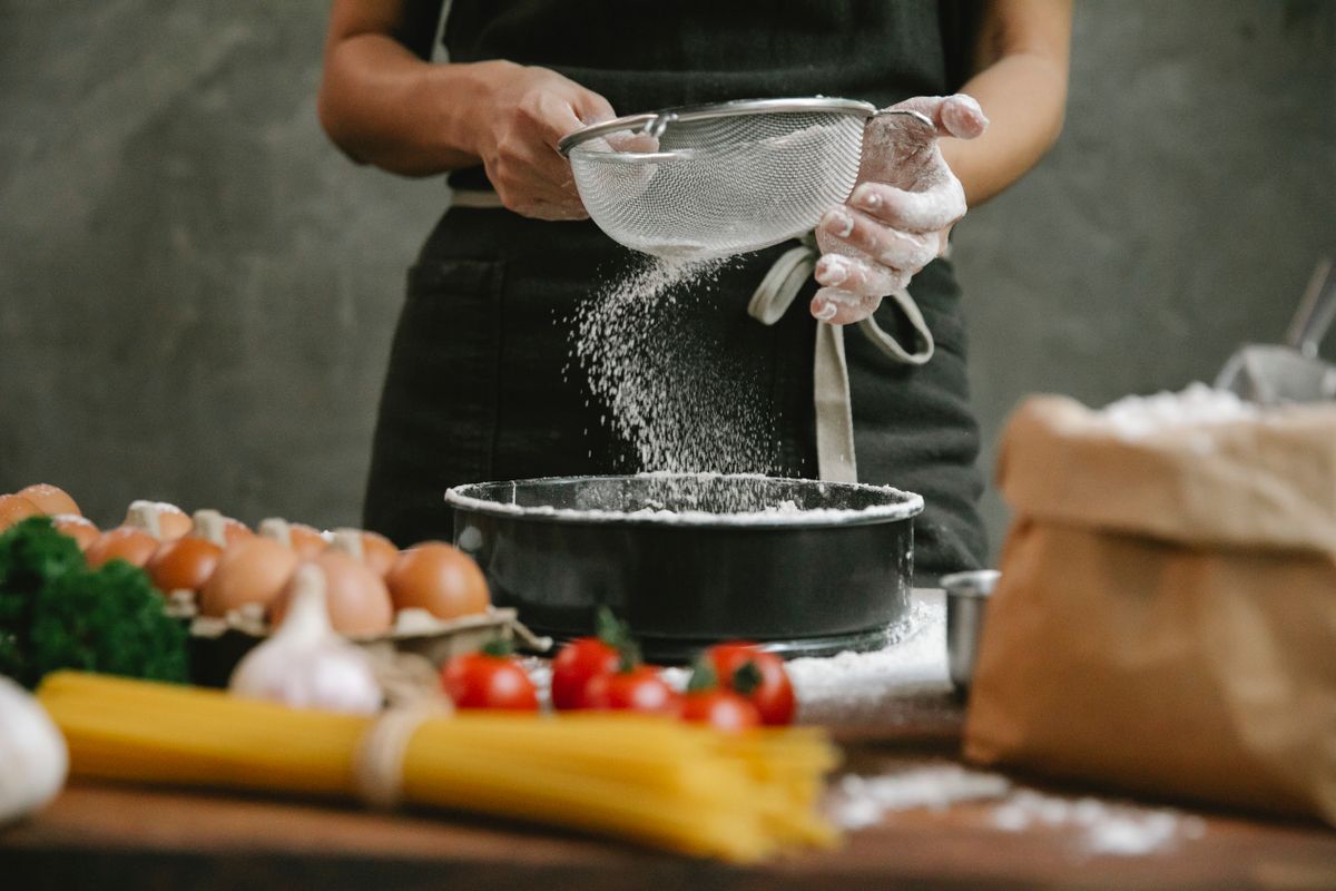 Crop anonymous chef adding flour to baking dish while making meal with eggs cherry tomatoes and spaghetti