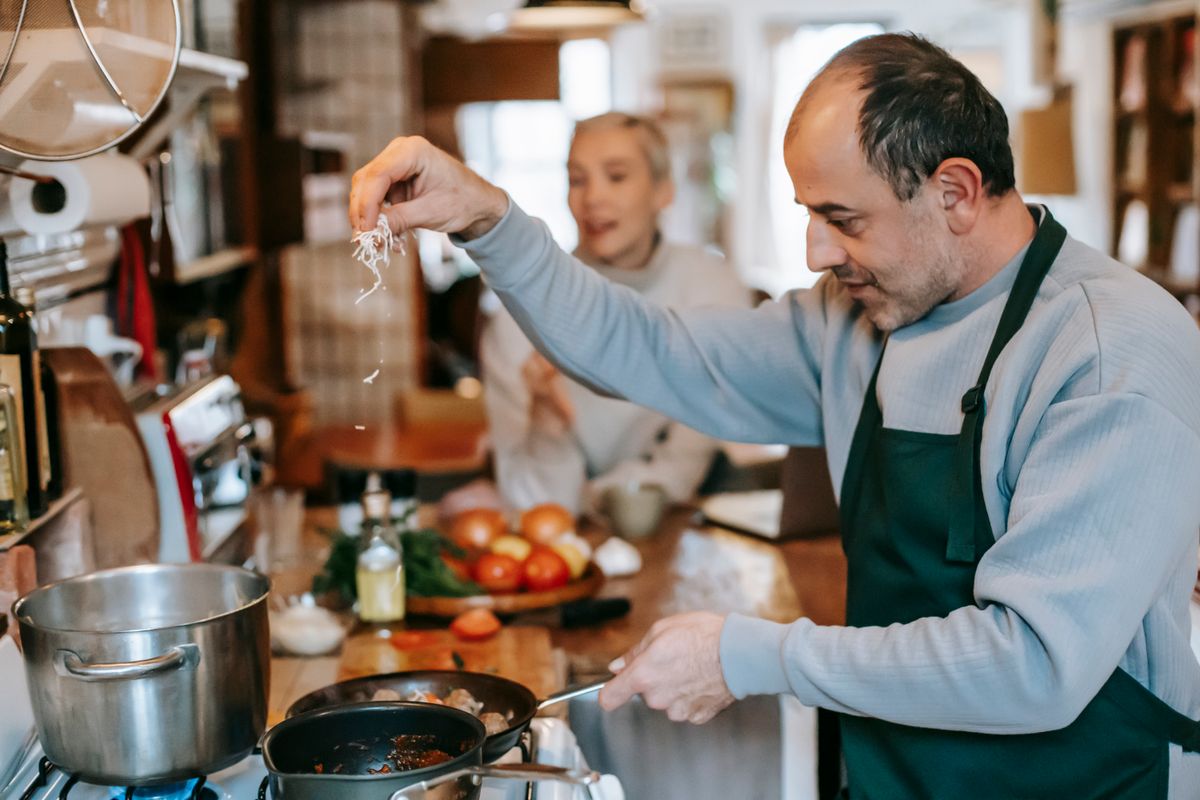 Focused male in apron adding ingredient into frying pan heating on gas stove while cooking meal for woman