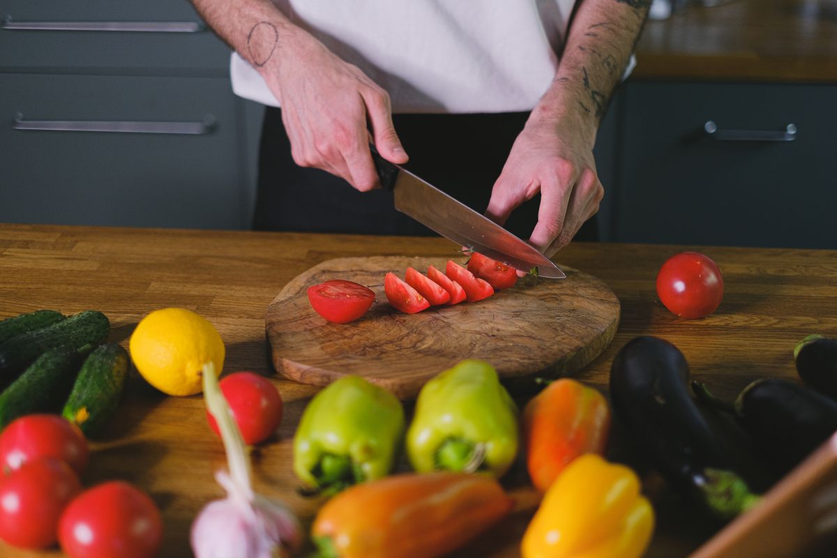 A person slicing tomatoes on a wooden chopping board with various fresh vegetables on a kitchen table.