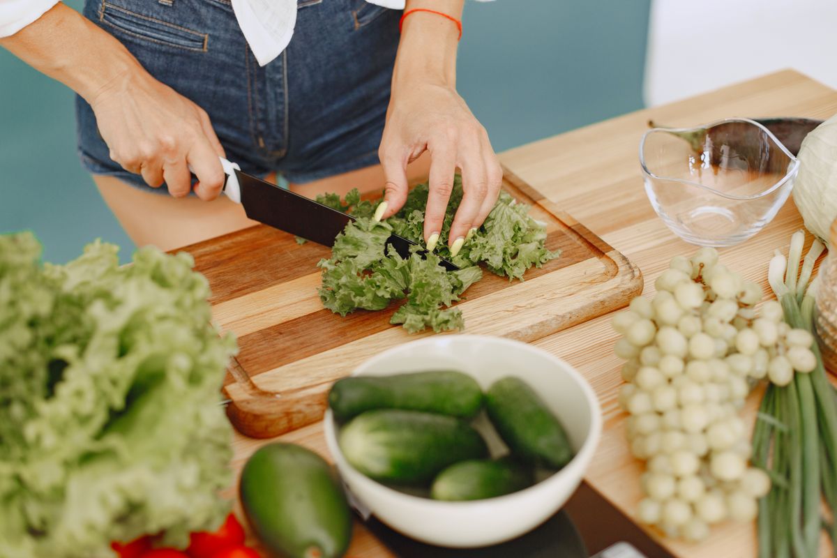 Hands chopping leafy greens on a wooden board with fresh vegetables for a healthy meal preparation.