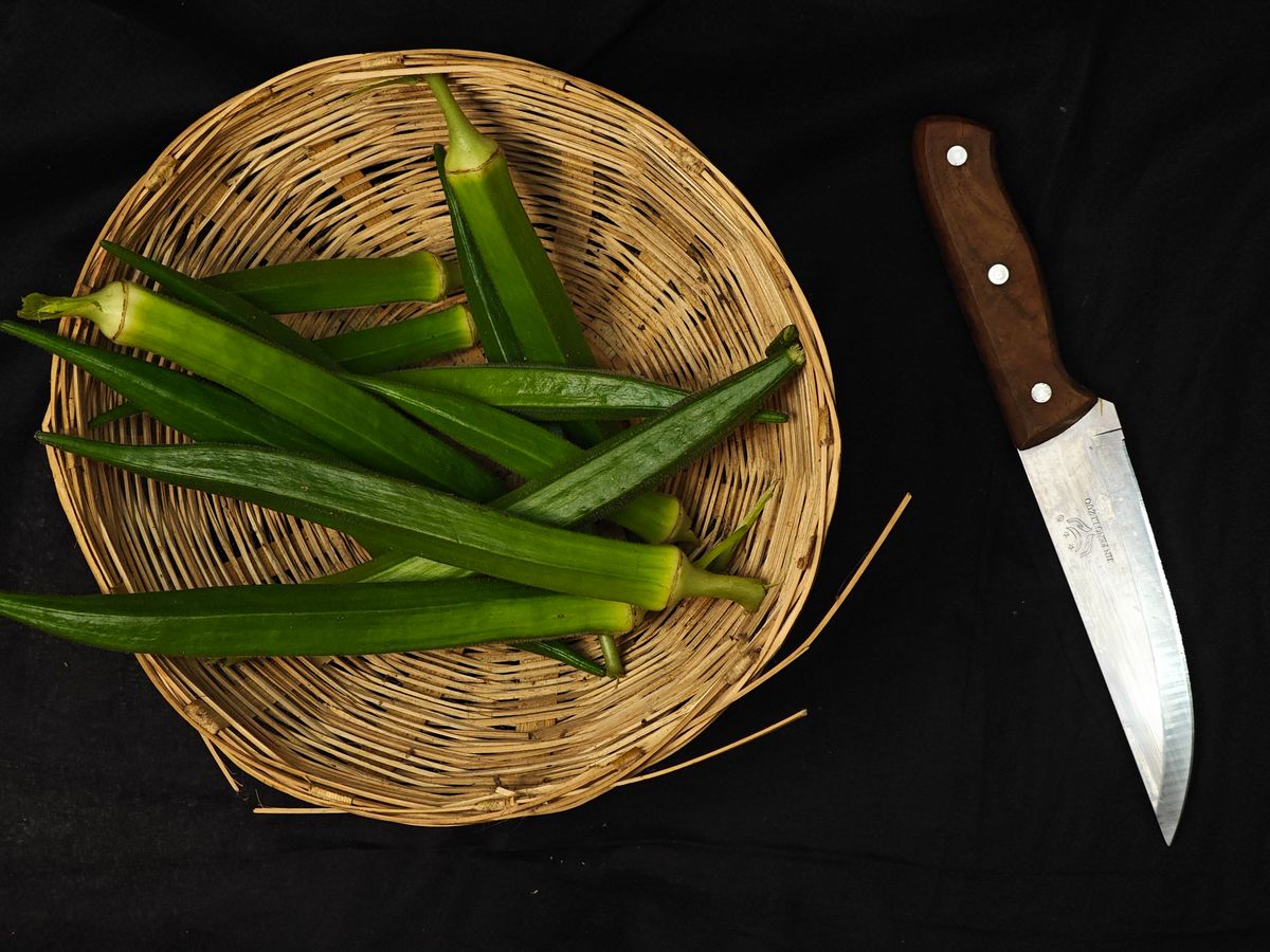Top view of fresh okra in a woven basket beside a wooden-handled kitchen knife.