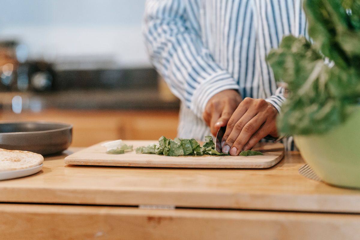 Crop anonymous ethnic male in striped shirt cutting fresh spinach on chopping board while cooking at home