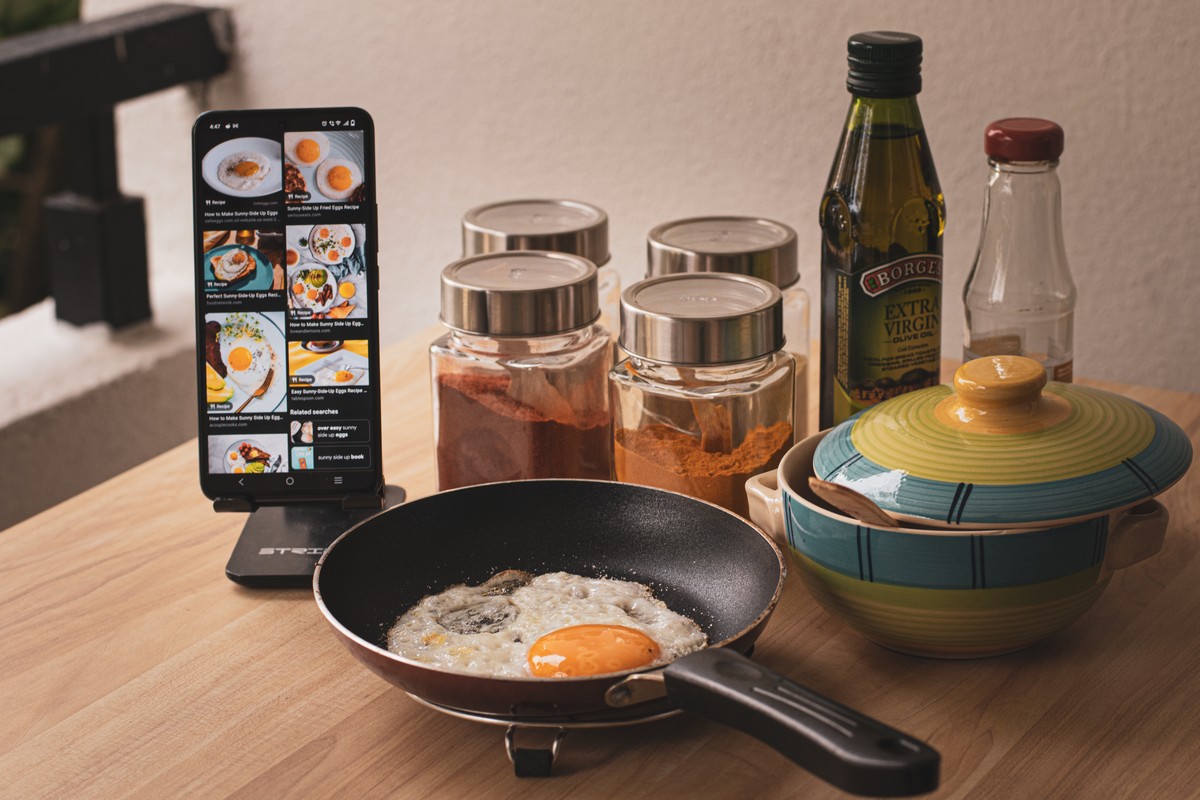 Fried egg cooking with smartphone recipe guidance, spices and ingredients on a kitchen counter.