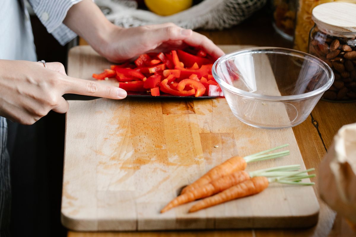 Anonymous female cook cutting red bell pepper on chopping board with carrots and bowl while preparing lunch in kitchen at home
