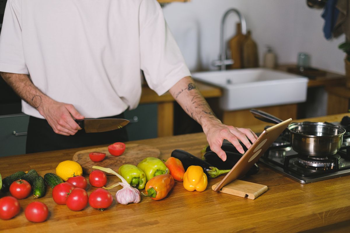 Adult man preparing vegetables using a knife and tablet in a contemporary kitchen setting.